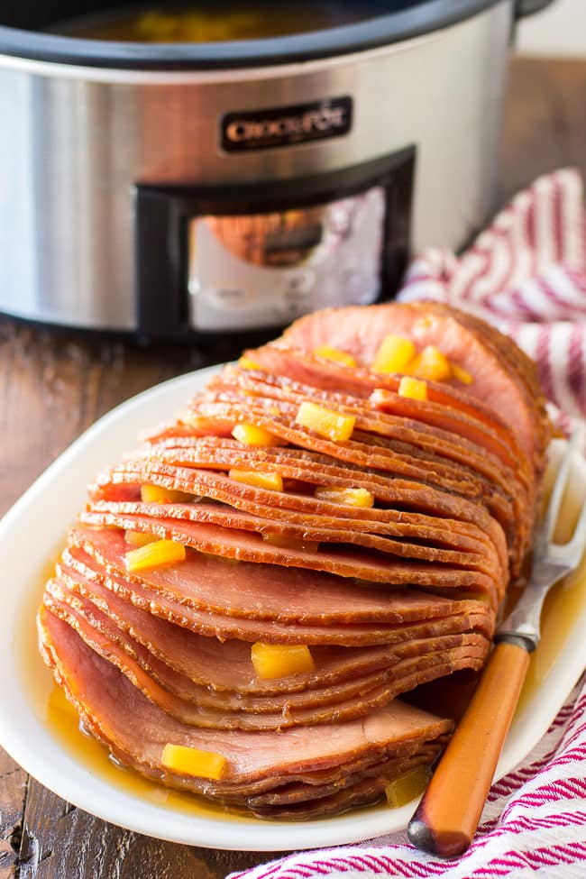 Juicy cooked Pork garnished with pineapple wedges between the sliced Pork, the crockpot is in the background, it is silver trimmed in black.